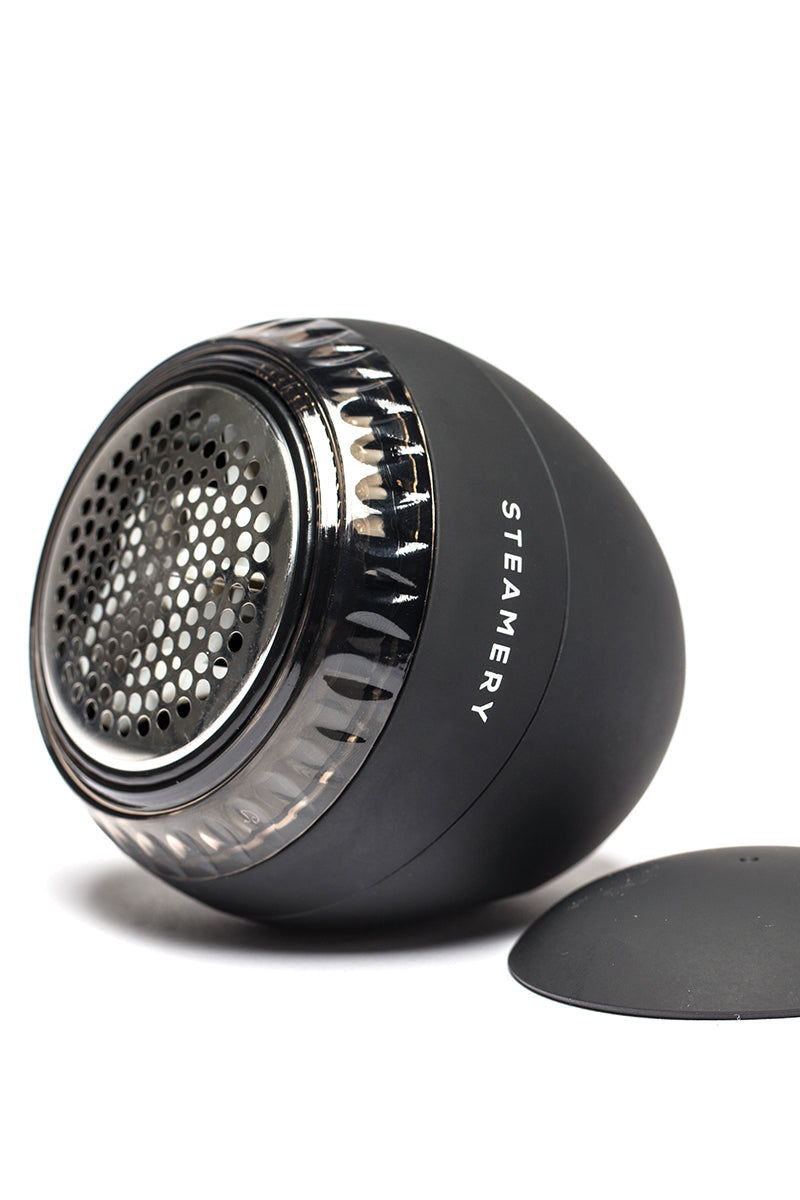 Eine Nahaufnahme eines schwarzen Steamery-Fusselentferners mit abgenommener Kappe zeigt die kleinen Löcher des Metallscherkopfes. Das Gerät ist seitlich abgewinkelt und trägt den Markennamen Steamery senkrecht auf der Seite.