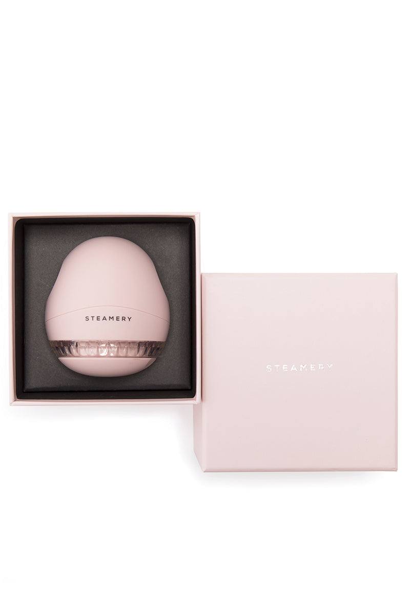 Ein blassrosa Steamery-Rasierapparat befindet sich in einer offenen, passenden rosa Schachtel mit einem durchsichtigen Deckel, auf dem die Klingen zu sehen sind. Auf dem rosa Schachteldeckel daneben steht "STEAMERY" in weißer Schrift. Der Hintergrund ist weiß.