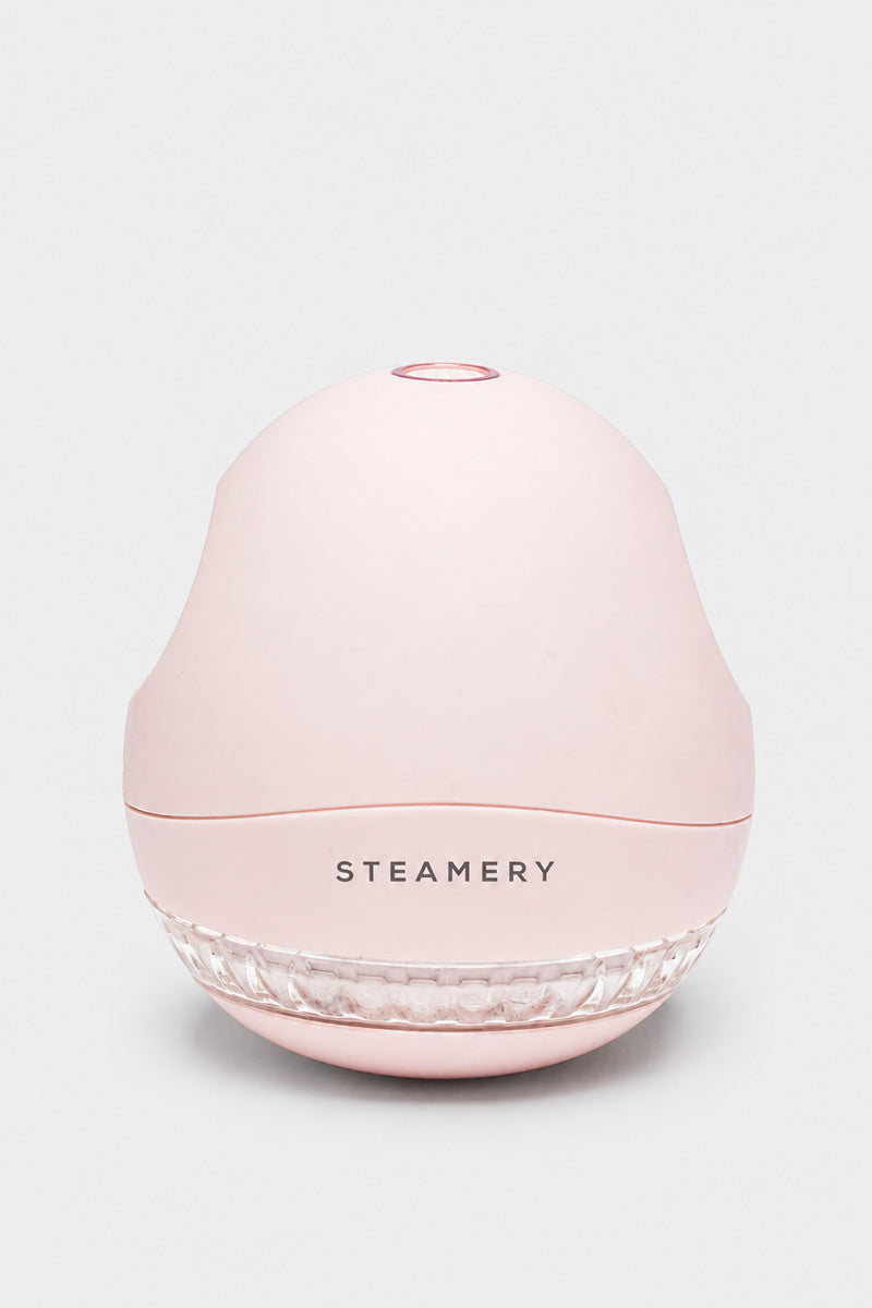 Ein hellrosa, kuppelförmiger Steamer mit der Aufschrift „STEAMERY“ mittig auf der Vorderseite. Der Sockel erinnert an das schlanke Design der Pilo 1 Fusselrasierer-Serie und weist einen durchsichtigen, geriffelten Bereich auf. Der Hintergrund ist einfarbig hellgrau, und das Produkt ist nach vorne gerichtet abgebildet.