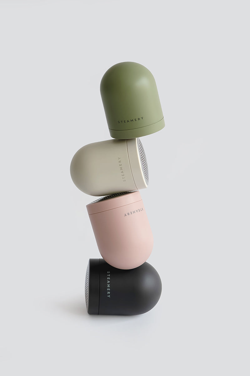 Vier runde, kuppelförmige Steamery Pilo 2-Rasierer in Grün, Beige, Pink und Schwarz sind vertikal vor einem hellgrauen Hintergrund gestapelt. Jedes Gerät ist angewinkelt, um eine minimalistische und moderne Komposition zu schaffen.