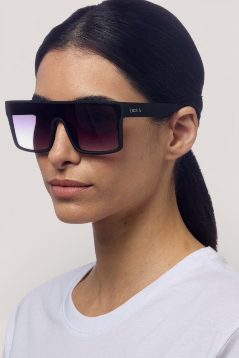 Eine Frau mit glattem, zurückgebundenem dunklem Haar trägt eine übergroße, rechteckige Sonnenbrille von Okkia mit Verlaufsgläsern und "OKKIA" auf dem Rahmen, ein schlichtes weißes Oberteil, heller Hintergrund, neutraler Ausdruck. Inklusive UV 400 Schutz.