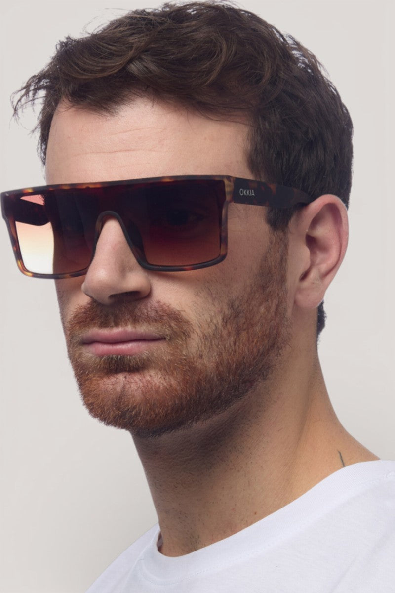 Ein Mann mit kurzen braunen Haaren und einem Bart trägt eine übergroße, rechteckige Sonnenbrille mit UV 400 Schutz von Okkia und ein weißes T-Shirt. Er blickt leicht zur Seite, mit neutralem Gesichtsausdruck, vor einem hellgrauen Hintergrund.