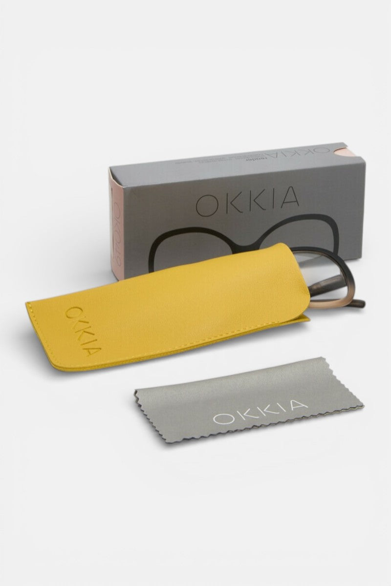 Eine Okkia-Lesebrille im Schmetterlingsdesign, teilweise in einem gelben Etui, liegt vor einer grauen Okkia-Box mit einer Brillengrafik. In der Nähe befindet sich ein graues Putztuch mit dem Okkia-Logo, alles auf weißem Hintergrund.