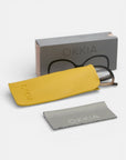 Eine Okkia-Lesebrille im Schmetterlingsdesign, teilweise in einem gelben Etui, liegt vor einer grauen Okkia-Box mit einer Brillengrafik. In der Nähe befindet sich ein graues Putztuch mit dem Okkia-Logo, alles auf weißem Hintergrund.