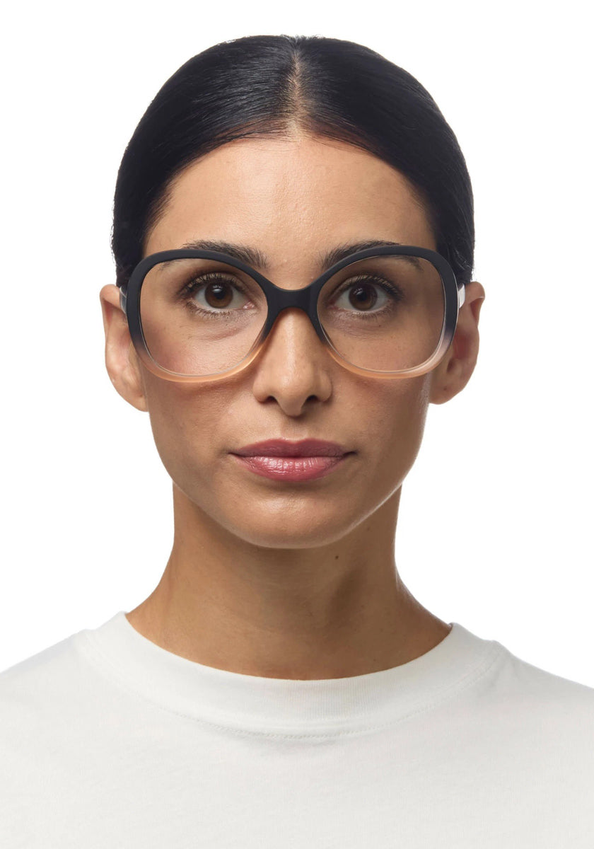 A woman with dark hair pulled back, wearing large black-rimmed Okkia Lesebrille and a white crew-neck shirt, looks directly at the camera against a plain white background. Her expression is neutral.