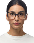A woman with dark hair pulled back, wearing large black-rimmed Okkia Lesebrille and a white crew-neck shirt, looks directly at the camera against a plain white background. Her expression is neutral.