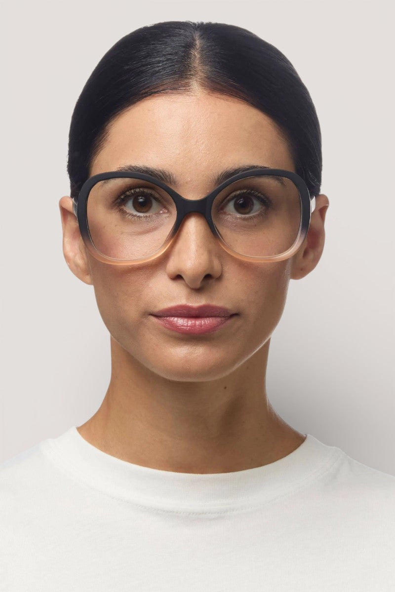 Eine Frau mit mittlerer Hautfarbe und glattem, dunklem Haar, das in der Mitte gescheitelt und nach hinten gezogen ist, trägt große, runde Okkia Lesebrille mit leichtem Farbverlauf und Soft-Touch-Oberfläche. Sie hat einen neutralen Ausdruck und trägt ein schlichtes weißes Oberteil vor einem hellgrauen Hintergrund.