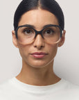 Eine Frau mit mittlerer Hautfarbe und glattem, dunklem Haar, das in der Mitte gescheitelt und nach hinten gezogen ist, trägt große, runde Okkia Lesebrille mit leichtem Farbverlauf und Soft-Touch-Oberfläche. Sie hat einen neutralen Ausdruck und trägt ein schlichtes weißes Oberteil vor einem hellgrauen Hintergrund.