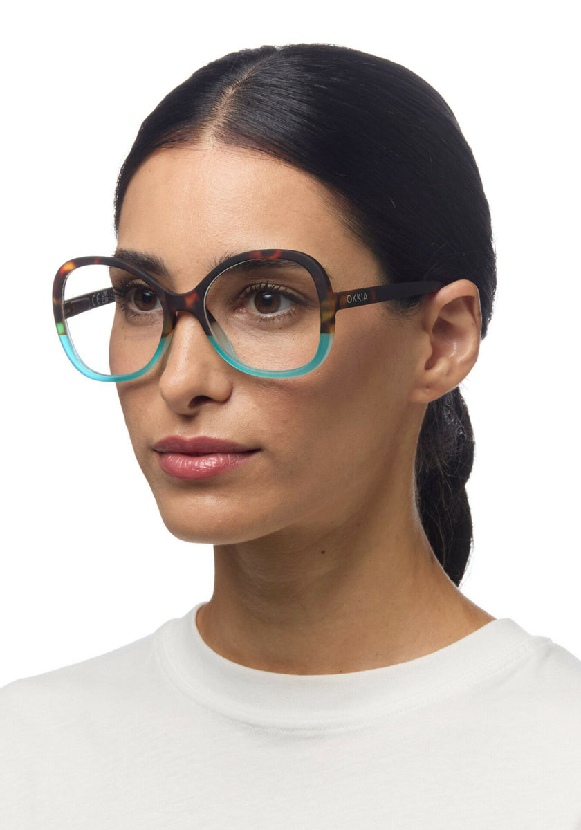 A woman with straight dark hair pulled back wears large, round Okkia Lesebrille featuring a multicolored frame—black on top, blue below, with red and yellow accents. She has a calm expression in a plain white top against a white background.