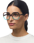 A woman with straight dark hair pulled back wears large, round Okkia Lesebrille featuring a multicolored frame—black on top, blue below, with red and yellow accents. She has a calm expression in a plain white top against a white background.