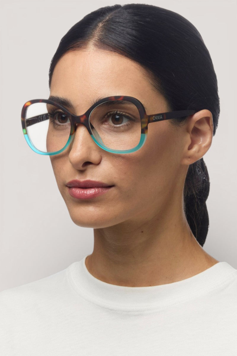 Eine Frau mit glattem, zurückgebundenem dunklem Haar trägt eine große, runde Okkia Lesebrille mit einem schildpattfarbenen und braunen Schmetterlingsmuster. Sie hat helle Haut, natürliches Make-up und ein schlichtes weißes Oberteil vor einem neutralen Hintergrund.