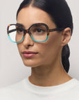 Eine Frau mit glattem, zurückgebundenem dunklem Haar trägt eine große, runde Okkia Lesebrille mit einem schildpattfarbenen und braunen Schmetterlingsmuster. Sie hat helle Haut, natürliches Make-up und ein schlichtes weißes Oberteil vor einem neutralen Hintergrund.