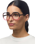 A woman with long dark hair tied back wears stylish Okkia reading glasses featuring large, round, two-tone frames in dark and pink. She has a neutral expression and is wearing a white top against a plain white background.
