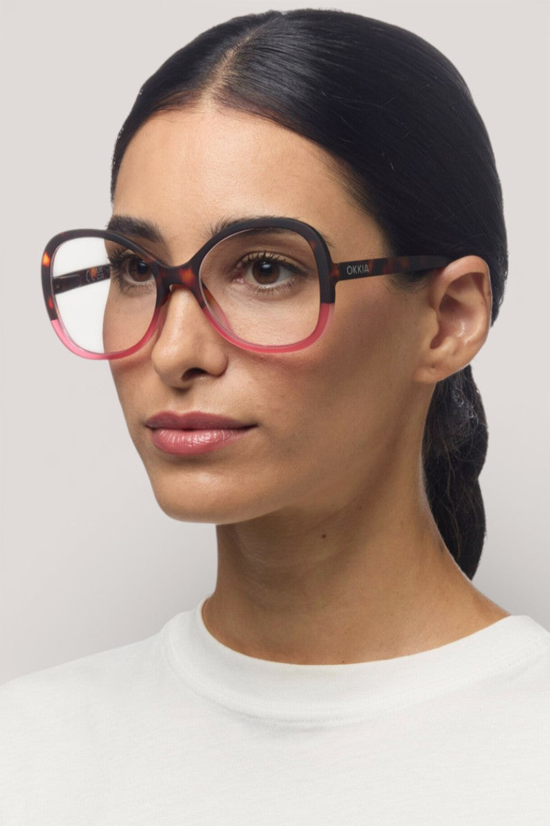 Eine Frau mit glattem, dunklem Haar trägt eine große, runde Okkia Lesebrille mit einem schwarz-rosa Schmetterlingsrahmen. Sie hat einen neutralen Ausdruck, ist weiß gekleidet und steht vor einem hellgrauen Hintergrund.