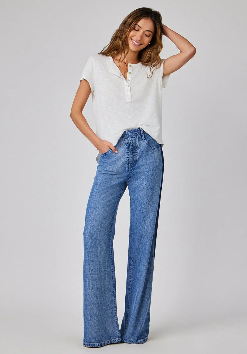 A woman with long, wavy hair smiles against a light background, wearing a Bella Dahl short-sleeve white henley shirt tucked into high-waist hose in denimoptik. One hand is in her pocket, and the other rests on her head.