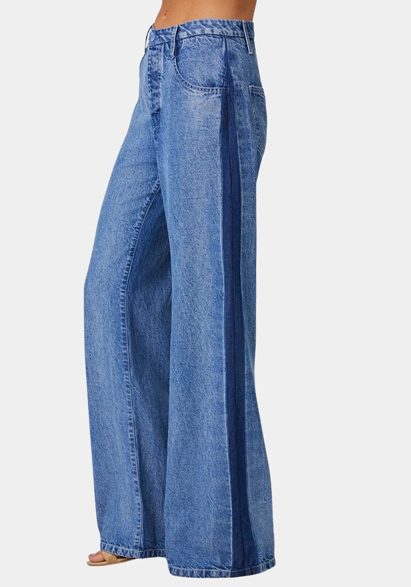 A person models Bella Dahl high-waisted, wide-leg blue jeans in denim look with a darker stripe on the side seam, visible stitching, front pockets, and a relaxed fit. The model stands sideways against a light gray backdrop.