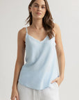 A woman with long brown hair wears a light blue Bella Dahl Lyocell Trägertop featuring frayed neckline and thin straps, paired with loose white pants. She stands against a plain background, looking relaxed with one hand in her pocket.
