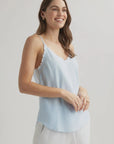A woman with long brown hair smiles while wearing a light blue Bella Dahl Lyocell Trägertop with frayed edges and light pants, standing against a plain, light background, facing left with her hands loosely clasped.