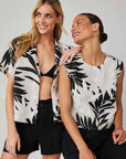 Two women smile. One wears black shorts, a black bikini top, and an open Bella Dahl Palmen-Print Leinenmix Hemdkragen blouse. The other sports a sleeveless white top with matching leafy print and black pants. Both have long hair; one down, one in a bun.
