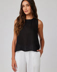 A woman with long wavy brown hair models a Bella Dahl sleeveless linen top and loose white pants, standing against a plain light gray background, facing forward with a relaxed expression and one hand in her pocket.