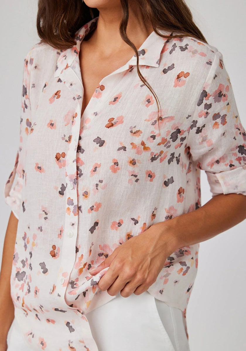 A woman models a light, semi-sheer Bella Dahl Leinenmix Bluse featuring a loose fit and delicate pink, orange, and gray Blumenprint. She holds the hem of the Hemdbluse with one hand, standing against a plain background.