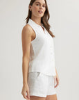 A woman with long brown hair models a sleeveless white button-up shirt with Hemdkragen and white shorts from Bella Dahl, standing against a plain light background. She faces slightly to the side, looking confidently at the camera.