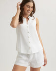A woman with long brown hair smiles while wearing a sleeveless, button-up white linen top with Hemdkragen and matching white shorts from Bella Dahl, touching her hair and standing against a plain light background.