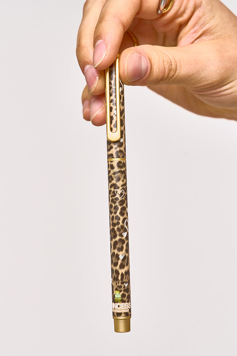 Eine Hand hält einen Princess Goes Hollywood Kugelschreiber mit Leopardenmuster und goldenen Akzenten vor einem schlichten hellen Hintergrund. Der Stift zeigt "INCESS" in der Nähe der Unterseite mit einer grünen und gelben Grafik, die Eleganz und Schreibkunst miteinander verbindet.
