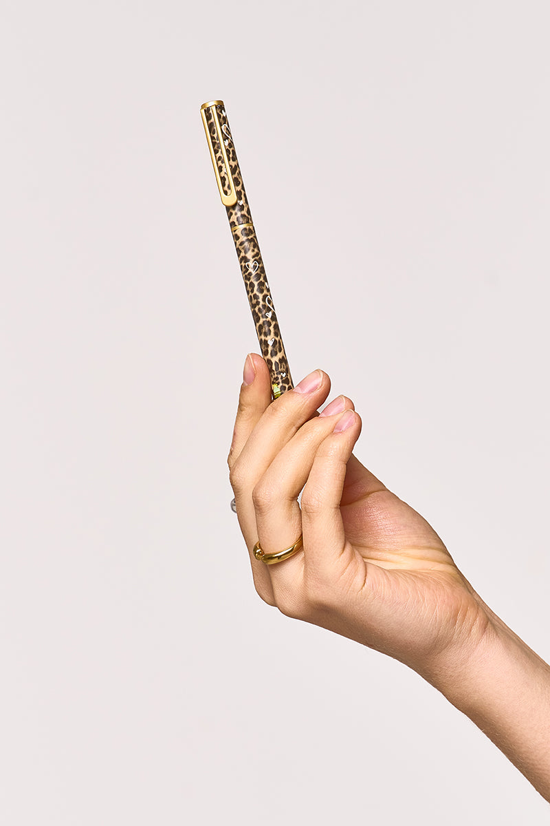 Eine Hand mit sorgfältig manikürten Nägeln und einem goldenen Ring hält einen Princess Goes Hollywood-Kugelschreiber mit Gold- und Leopardenmuster vor einem schlichten, hellen Hintergrund, der Eleganz ausstrahlt, während sich die Finger anmutig darum winden.