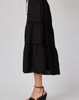 A person wears a black Leinen Midirock by Bella Dahl with Stufenschnitt, a drawstring waist, and raw hem, paired with black wedge sandals. The photo captures a side view against a plain light gray background.