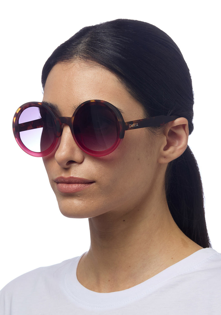 A woman with straight dark hair tied back wears large, round Okkia sunglasses with gradient pink-tinted lenses and tortoiseshell frames. She has a neutral expression and is dressed in a plain white shirt against a white background.