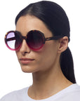 A woman with straight dark hair tied back wears large, round Okkia sunglasses with gradient pink-tinted lenses and tortoiseshell frames. She has a neutral expression and is dressed in a plain white shirt against a white background.