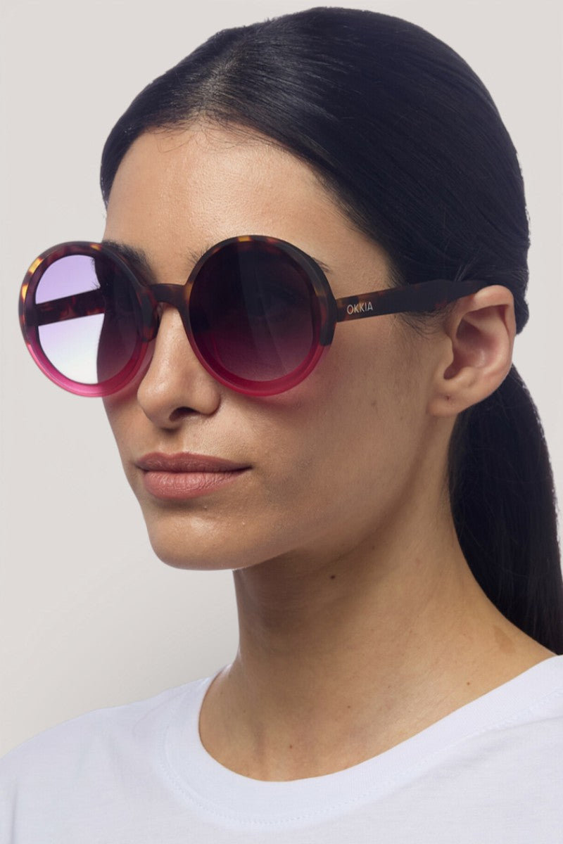 Eine Frau mit glattem, dunklem Haar trägt eine große, runde Sonnenbrille Okkia Oversize mit einem Gestell aus Schildpatt und rosafarbenem Farbverlauf und UV 400 Schutz. Sie hat einen neutralen Gesichtsausdruck und trägt ein weißes Oberteil vor einem hellgrauen Hintergrund.