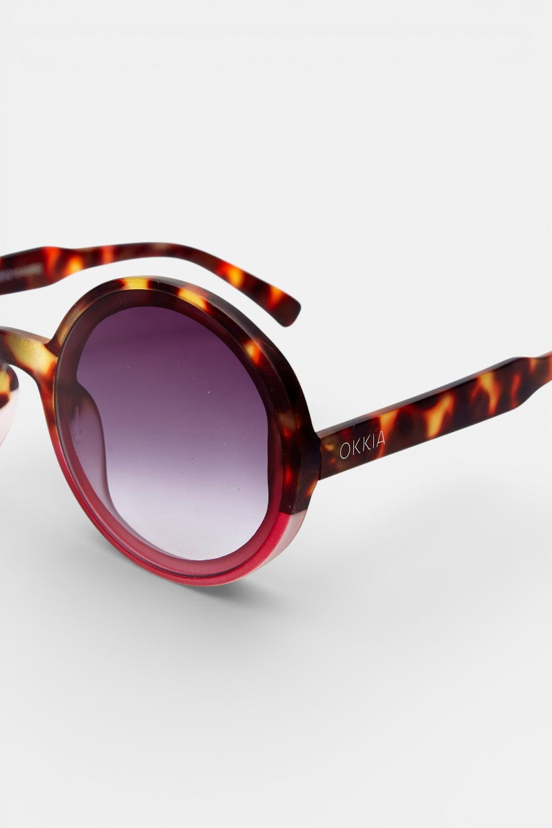 Nahaufnahme der runden Sonnenbrille Okkia mit schildpattfarbenem Rahmen und lila Verlaufsgläsern. Der Schriftzug "Okkia" ist in Weiß auf die Bügel gedruckt. Bietet UV-400-Schutz für optimale Sicherheit der Augen. Vor einem schlichten weißen Hintergrund.