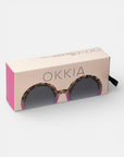 Eine rechteckige, beigefarbene Schachtel mit "Okkia" in fetten schwarzen Buchstaben auf der Oberseite präsentiert eine runde Sonnenbrille mit Schildpatt-Rahmen, UV-400-Schutz-Gläsern, rosafarbenen Seiten und einer schwarzen Zuglasche - ideal für einen stilvollen Augenschutz.