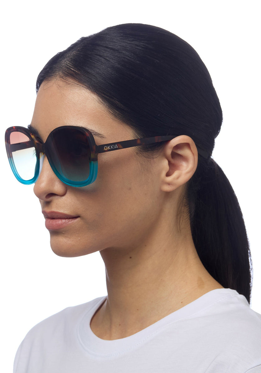 A woman with straight black hair in a low ponytail is shown in profile wearing Okkia gradient sunglasses—brown at the top and turquoise-blue at the bottom. She wears a white shirt against a plain white background.