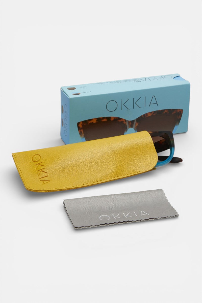 Eine Sonnenbrille von Okkia mit schildpattfarbenem Rahmen befindet sich teilweise in einem gelben Etui. Davor liegt ein graues Putztuch, und dahinter ist eine hellblaue Okkia-Schachtel mit einer Abbildung der Sonnenbrille im Cat-Eye-Design zu sehen.