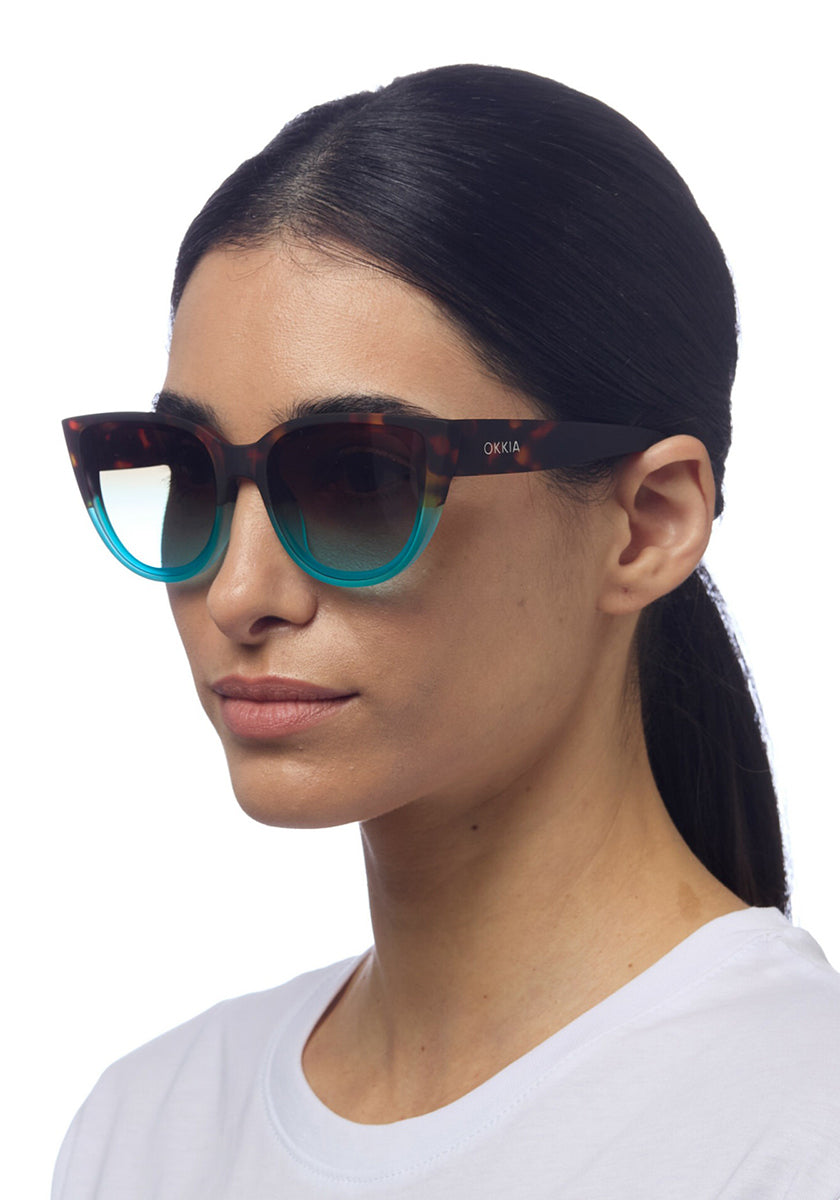 A woman with straight dark hair pulled back wears a white shirt and large Okkia cat-eye sunglasses that fade from tortoiseshell brown to turquoise blue, featuring the Okkia logo on the arms. The background is plain white.