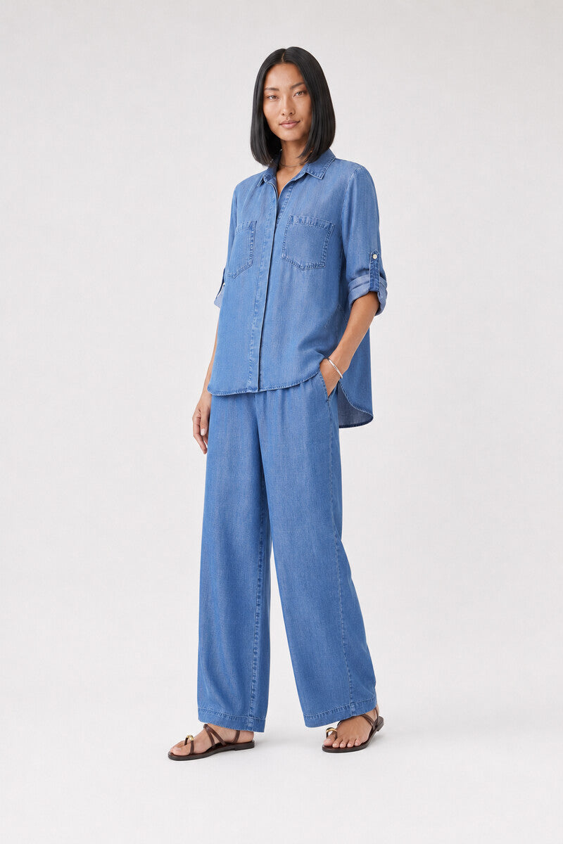Eine Person mit glattem, schulterlangem schwarzem Haar trägt eine locker sitzende, hellblaue Bella Dahl Damen-Hemdbluse aus Denim mit hochgekrempelten Ärmeln und dazu passende weit geschnittene Jeanshosen. Sie steht mit einer Hand in der Tasche vor einem schlichten hellen Hintergrund und trägt flache schwarze Sandalen.