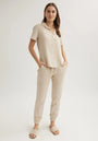 A woman with light brown hair, parted in the middle, stands against a plain background wearing a light beige Bella Dahl Lyocell Kurzarmbluse with Hemdkragen, matching drawstring jogger pants, and beige slide sandals. Her hands are in her pockets.