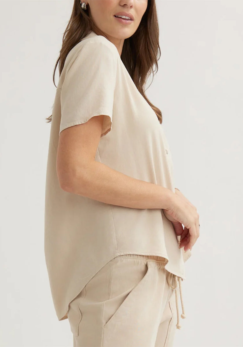 A woman in side profile wears a loose Bella Dahl Lyocell short-sleeve blouse with shirt collar and matching light beige pants. She has long brown hair, earrings, and lightly rests her hands near her waist against a plain background.