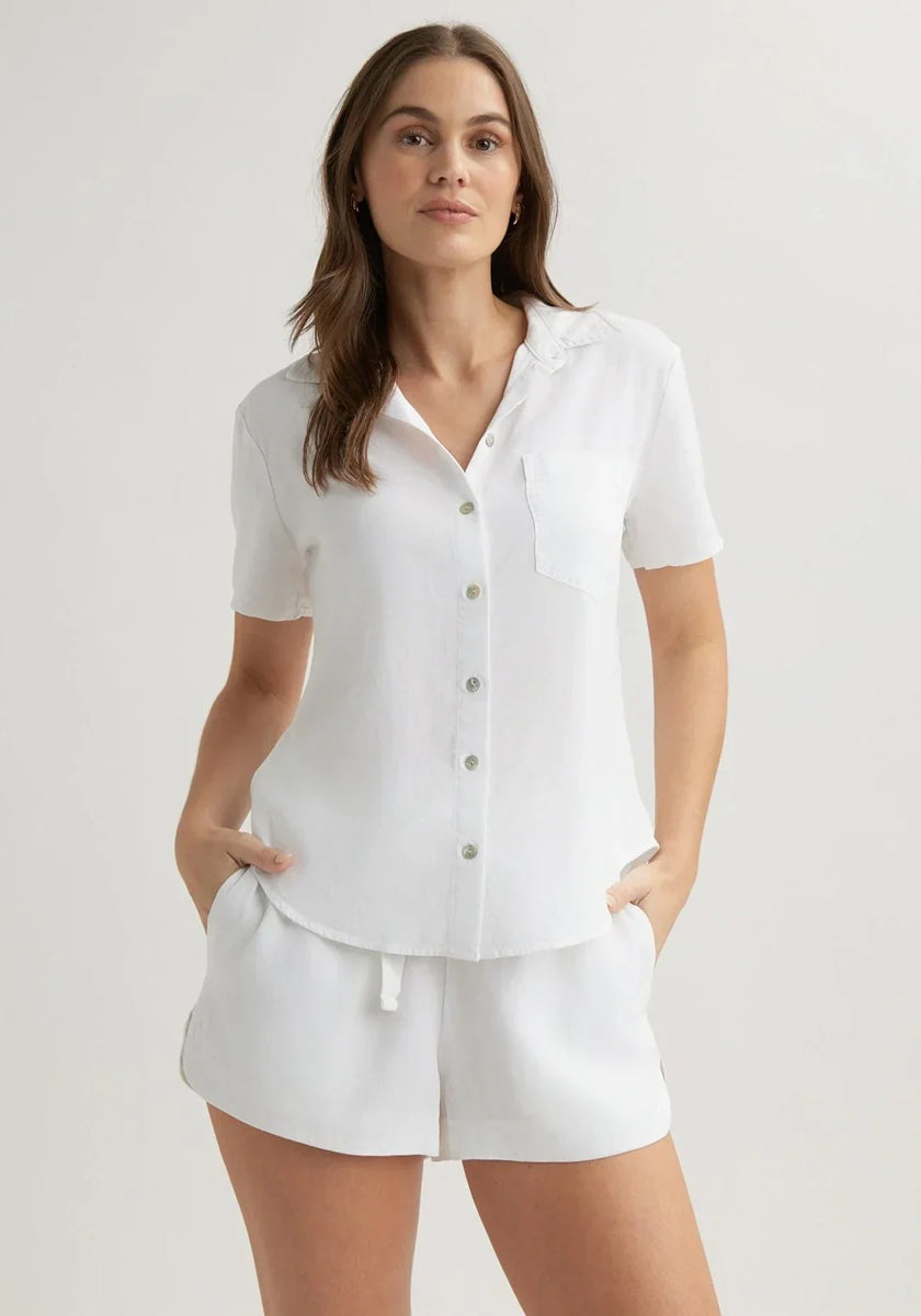 A woman with long brown hair wears a Bella Dahl kurzarmbluse with Hemdkragen and chest pocket, paired with matching white shorts. She stands confidently, hands in pockets, against a plain, light-colored background.