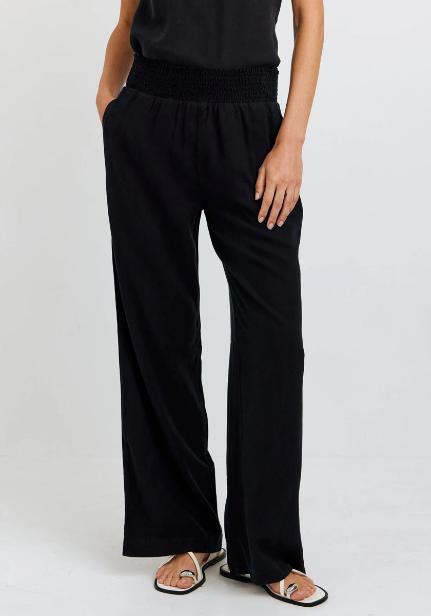 A person stands against a plain background wearing Bella Dahl weit geschnittene Hose in Denimoptik with a wide elastic waistband, paired with a black sleeveless top and white sandals. Their hands are in their pockets, head out of frame.