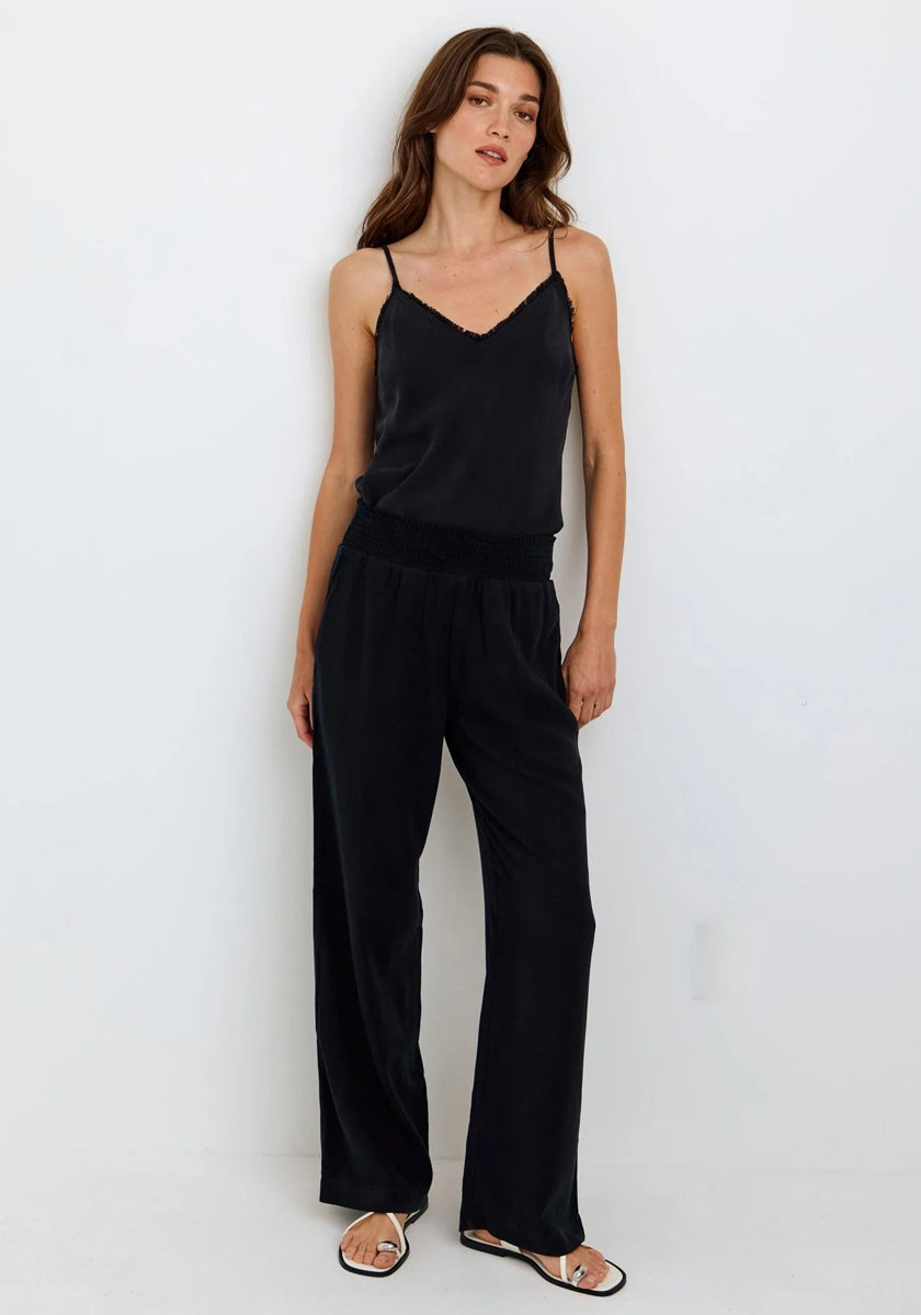 A woman with long brown hair and neutral makeup stands against a white wall, wearing a Bella Dahl sleeveless black V-neck camisole with lace trim and relaxed-fit wide-leg pants, paired with black flat sandals. She appears calm and relaxed.