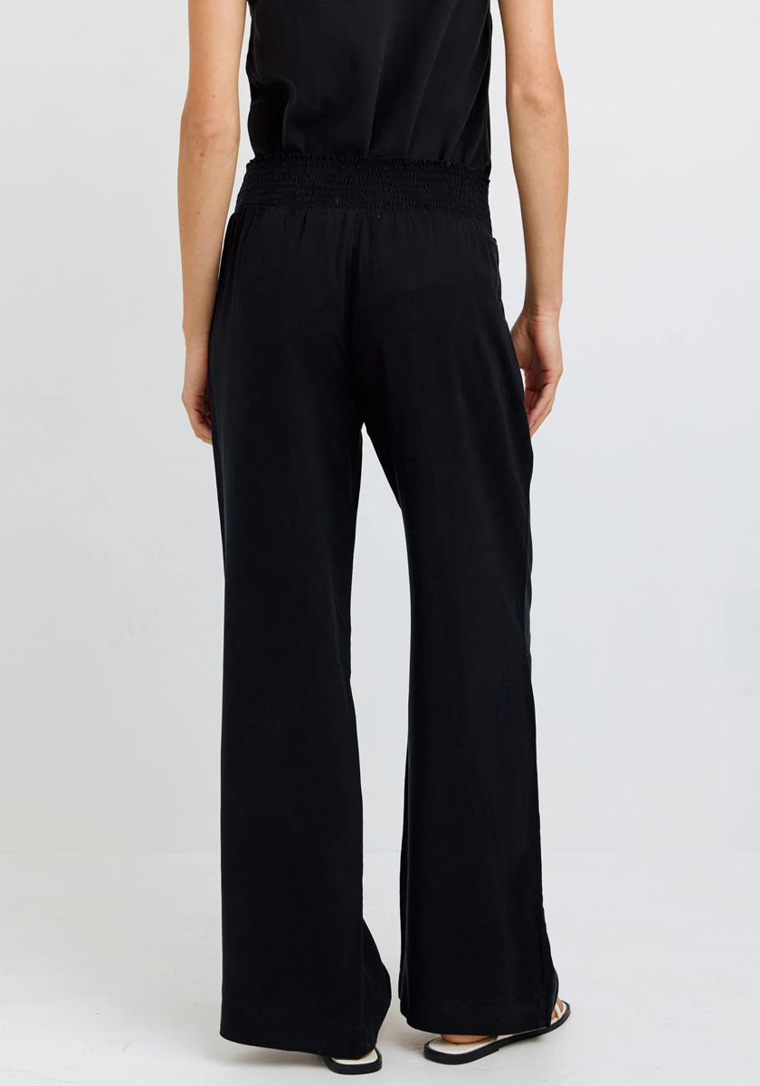 A person stands with their back to the camera, wearing wide-leg denim-look Lyocell pants with a smocked waistband by Bella Dahl, a sleeveless black top, and flat black sandals against a light background.