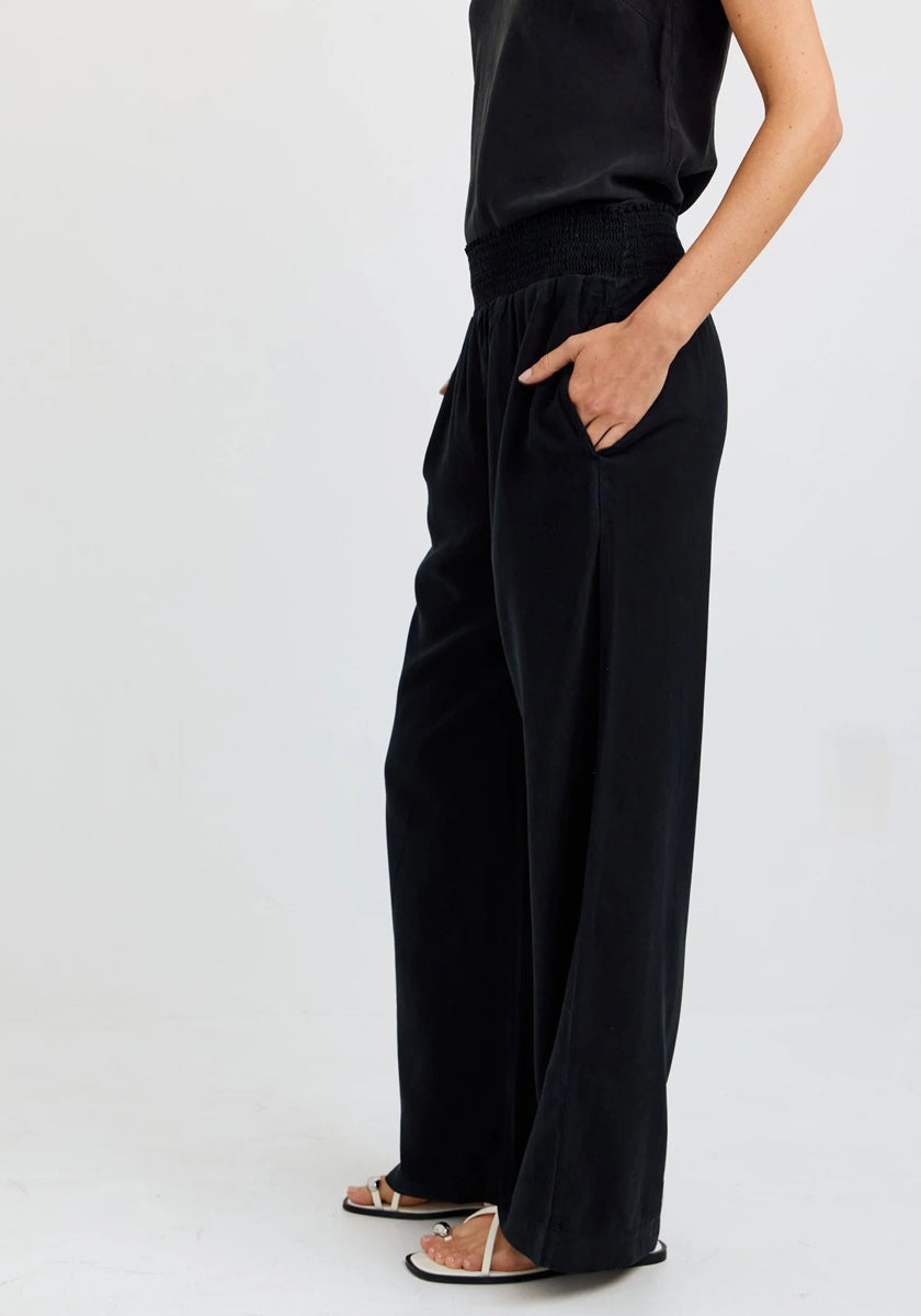 A person stands against a white background wearing Bella Dahls black wide-leg pants with an elastic waistband, a black sleeveless top, and flat white sandals. Their hands are in their pockets and their face is not visible.