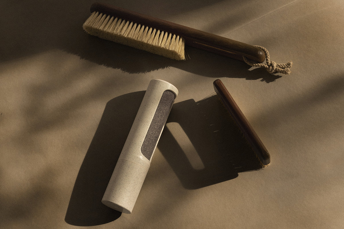 Die hölzerne Kleiderbürste mit cremefarbenen Borsten, der Holzkamm und der wiederverwendbare Fusselentferner mit Netzeinsatz von Steamery sind auf einer beigen Fläche ausgestellt, die bei warmem Tageslicht weiche Schatten wirft.