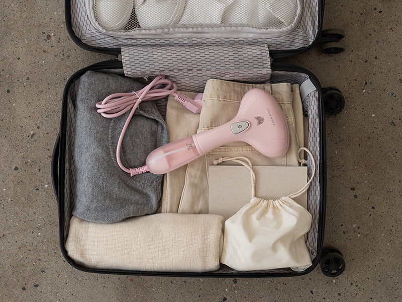 Ein offener Koffer auf einem gesprenkelten Boden, in dem sich ordentlich gefaltete beige, graue und cremefarbene Kleidungsstücke, ein rosa „Steamery“-Handdampfglätter mit Kabel zum Glätten von Kleidung, ein kleiner Beutel mit Kordelzug und ein beiger, rechteckiger Beutel befinden. Oben ist teilweise eine mit Netzstoff ausgekleidete Innentasche zu sehen.
