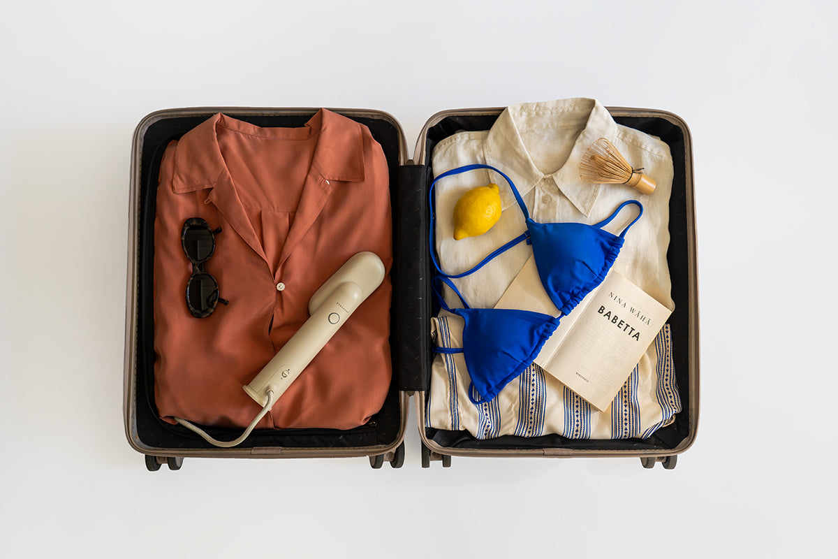 Ein offener Koffer mit ordentlich gepackten Gegenständen: links ein gefaltetes rostfarbenes Hemd, eine schwarze Sonnenbrille und ein tragbarer Kleidungsdampfer von Steamery; rechts ein weißes Hemd, ein Bambusbesen, eine Zitrone, ein blaues Bikinioberteil und das Buch Babetta.