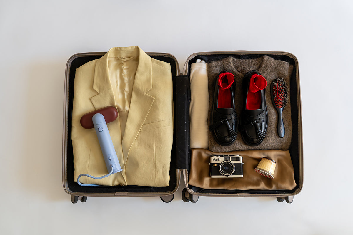 Ein offener Koffer auf weißem Hintergrund enthält einen gelben Blazer, einen dampfenden Reisesteamer, ein zusammengerolltes weißes Handtuch, schwarze Schuhe mit rotem Futter, einen braunen Pullover, eine Haarbürste, eine Kamera und eine goldene Tasse.